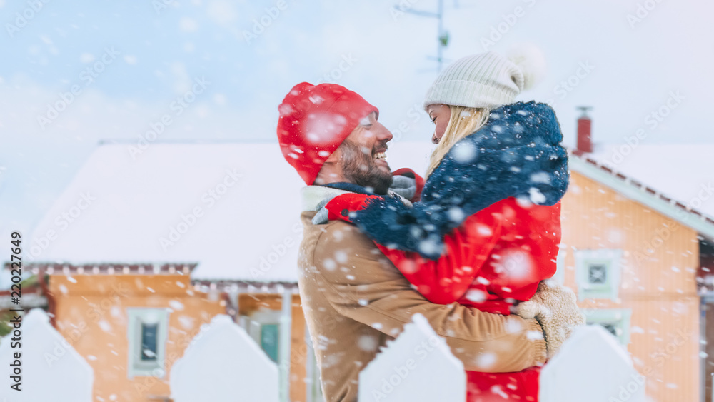 美丽的年轻情侣在他们田园风格的房子后院拥抱，而雪却浪漫地飘落。