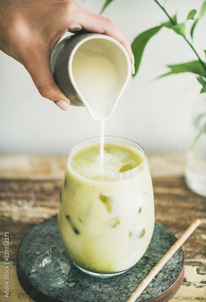 装在玻璃杯里的抹茶拿铁冰饮料，椰奶由女人的手从水罐里倒出来，白墙