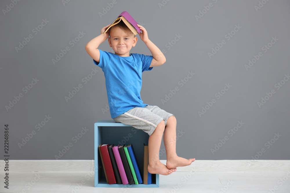 Cute little boy with books sitting on shelf near grey wall indoors