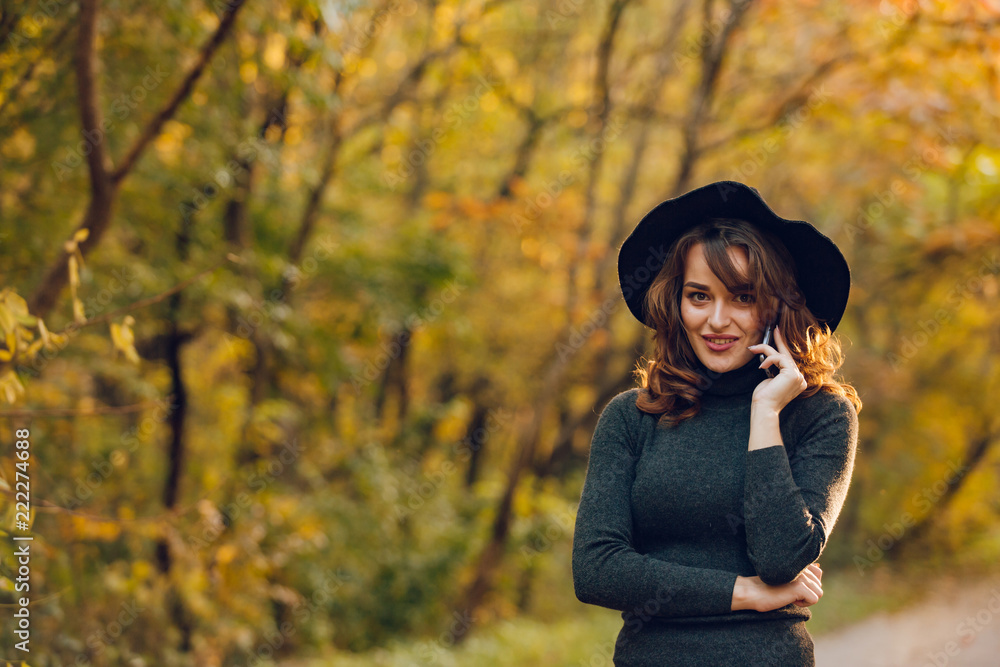 beautiful girl in a black hat holds a phone in her hand in the park in the autumn. Woman walk in the