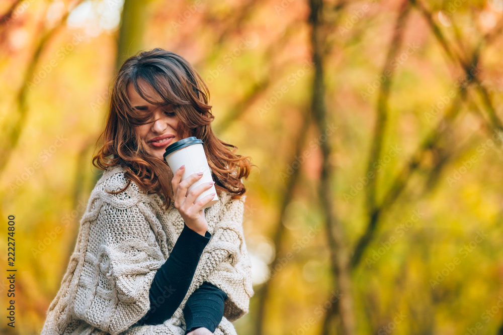 pretty girl with a cup of hot drink smiles in the forest in the autumn. Woman dressed in a warm swea