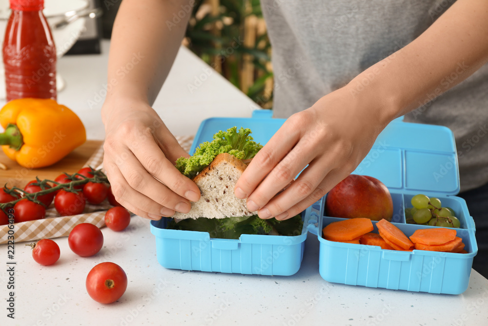 Mother putting food for schoolchild in lunch box at table, closeup