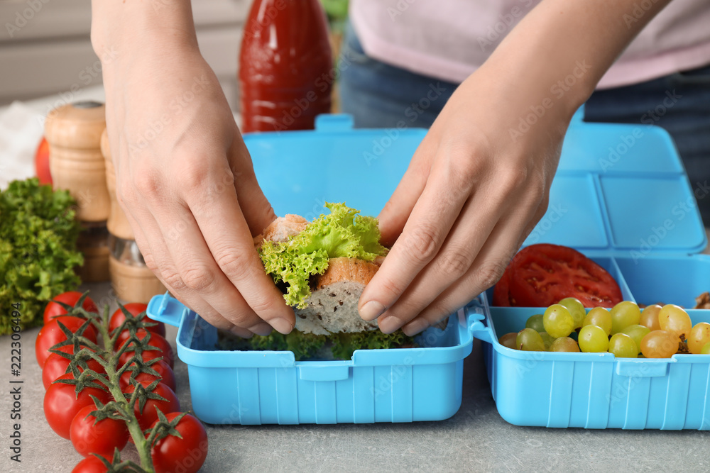 Mother putting food for schoolchild in lunch box at table, closeup