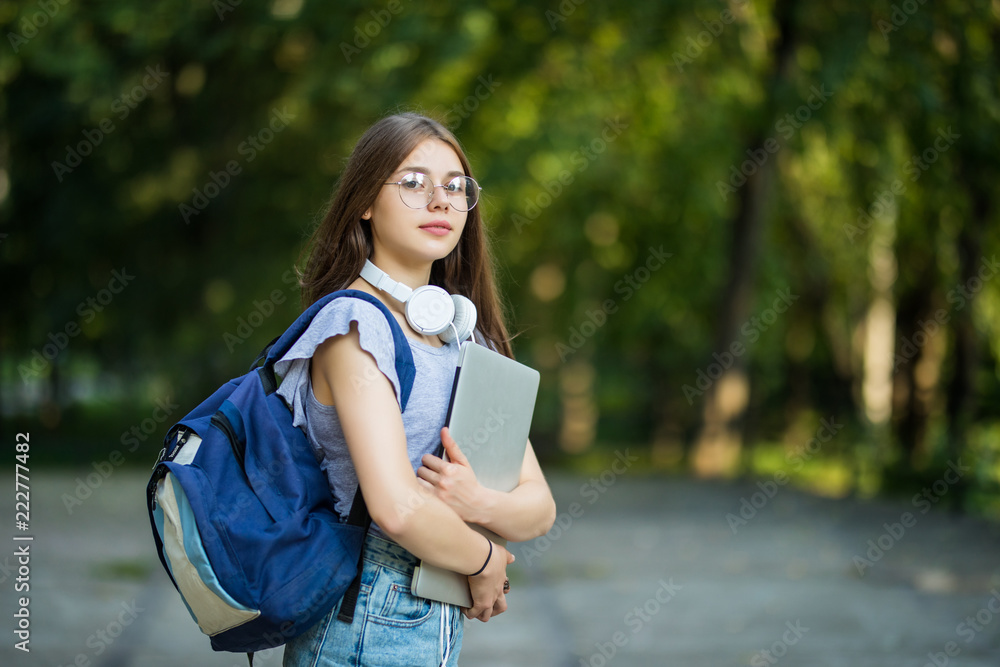 带着双肩包和笔记本站在公园里微笑的快乐迷人的年轻女性