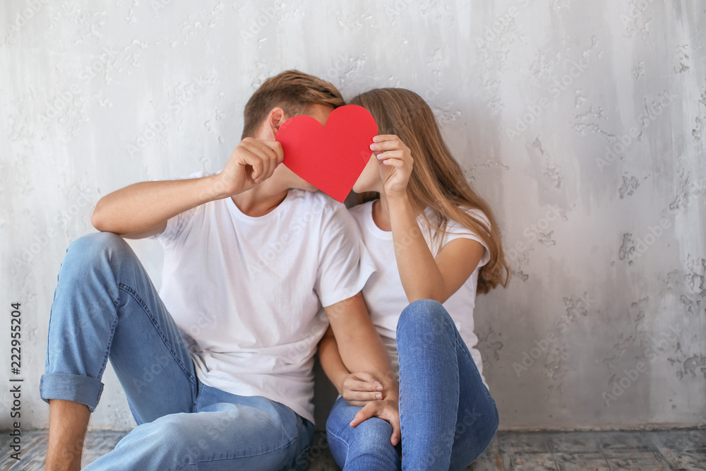 Happy young couple hiding faces behind red heart while sitting on floor at home