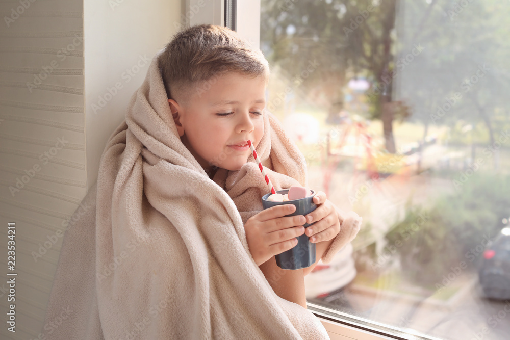 Cute little boy wrapped in warm plaid with hot chocolate in cup sitting on windowsill at home