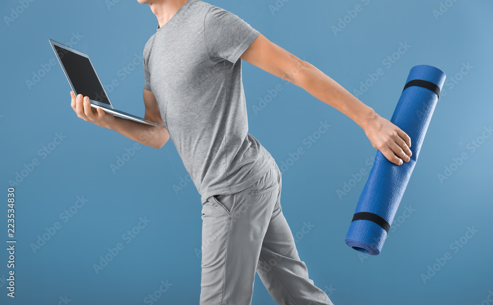 Young man with yoga mat and laptop running on color background. Concept of balance between rest and 