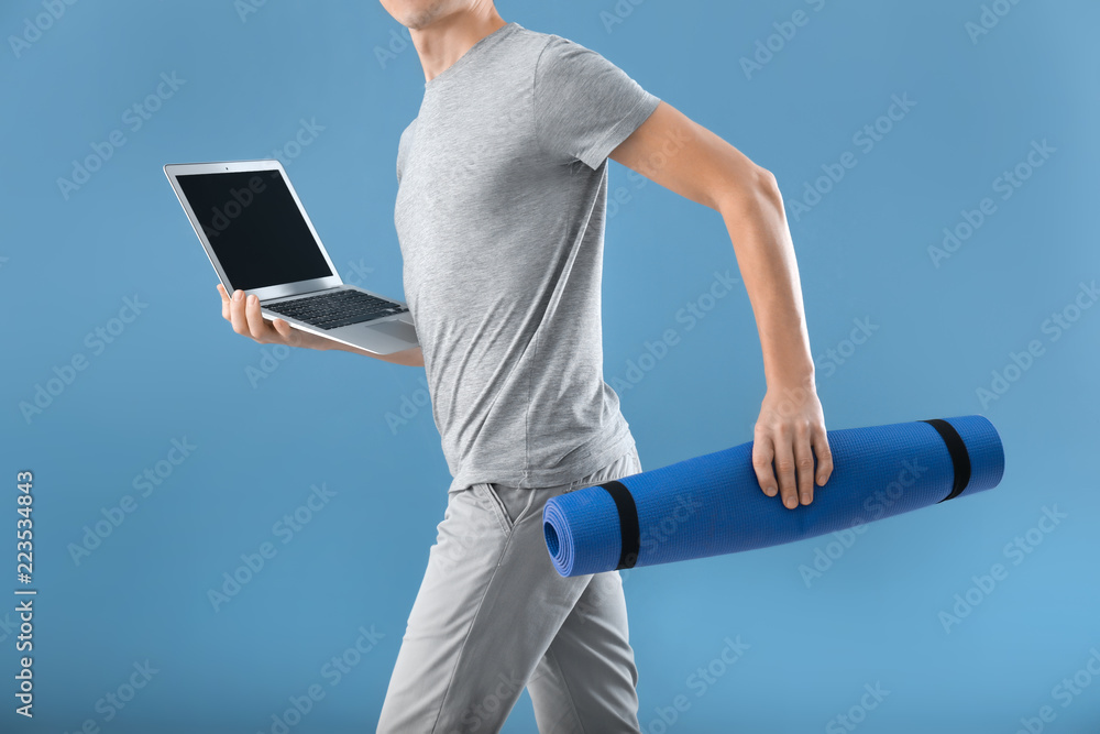 Young man with yoga mat and laptop running on color background. Concept of balance between rest and 