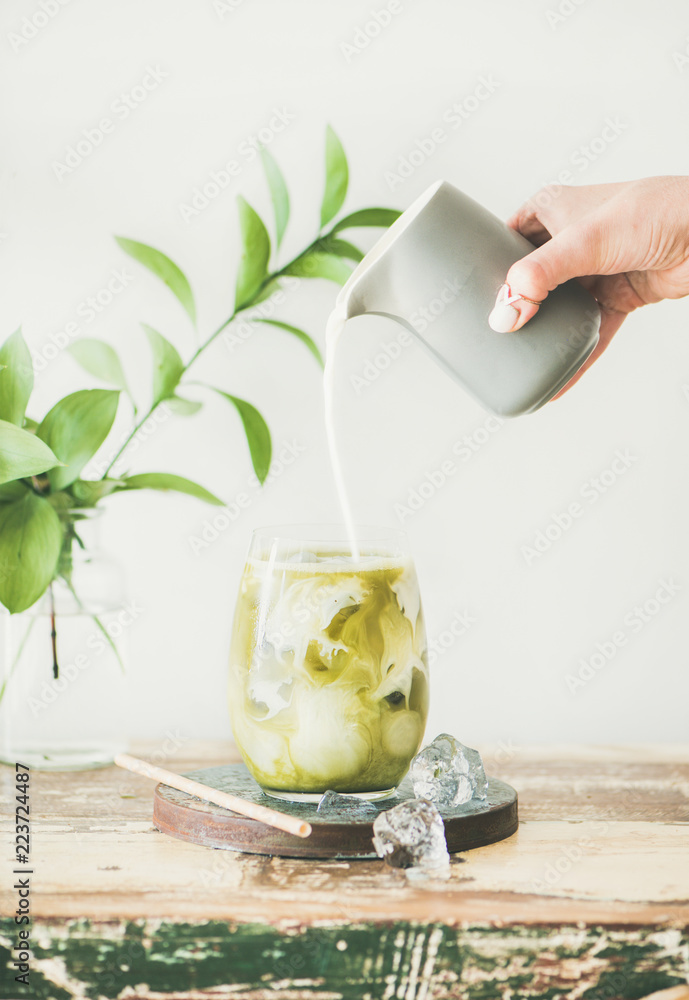 杯装冰镇抹茶拿铁饮料，用女性的手从水罐里倒入椰奶，白色墙壁