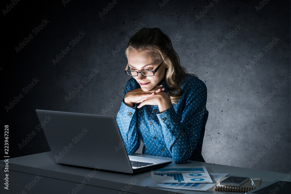 Attractive blonde working on laptop in dark office. Mixed media