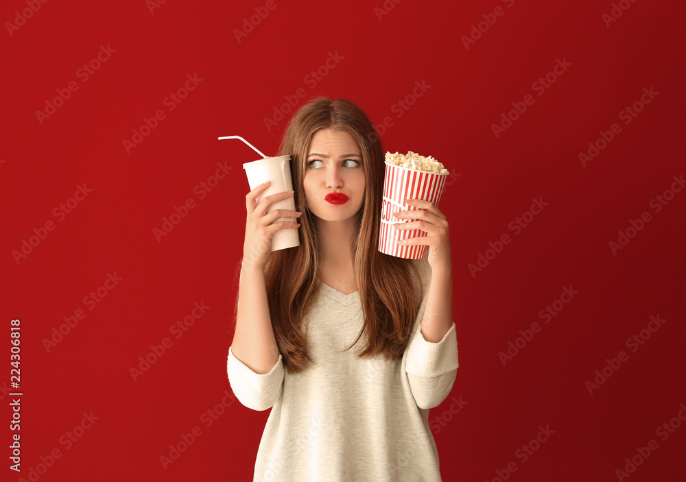 Emotional young woman with cup of popcorn and beverage on color background