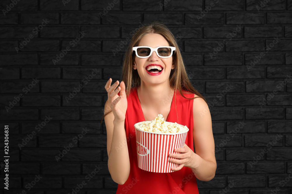 Beautiful young woman with cup of popcorn and 3D cinema glasses against dark brick wall