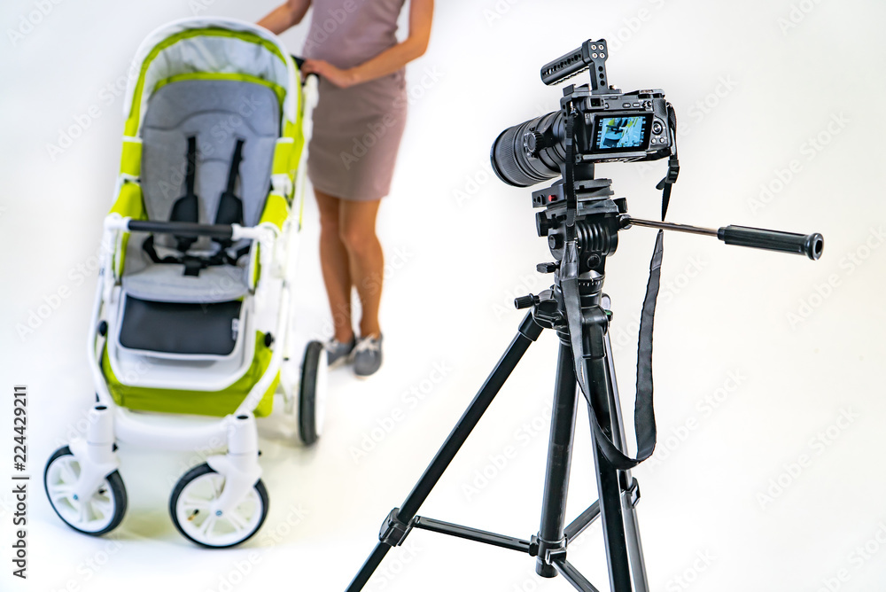 professional camera stands on a tripod and focuses on a woman in pink dress who holds stroller by he