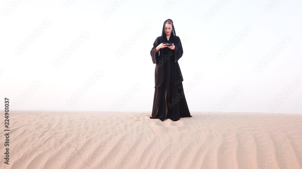 A woman in abaya (United Arab Emirates traditional dress) flying a drone on the dunes in the desert 