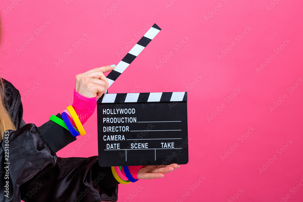 Woman in 1980s fashion holding a film movie slate board clipper on a pink background