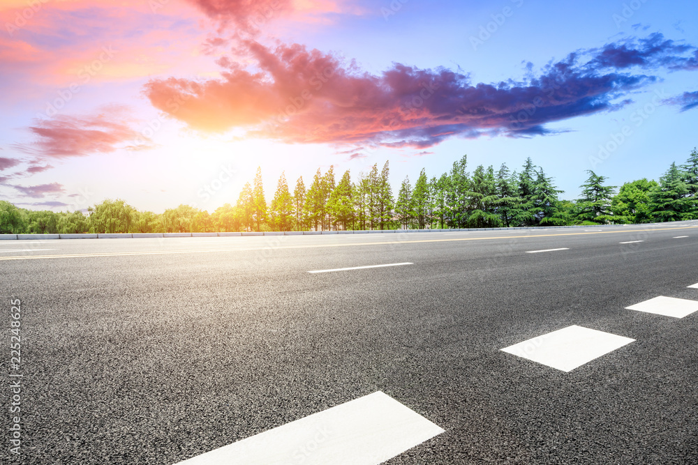 Empty asphalt road and green forest with colorful clouds at sunset