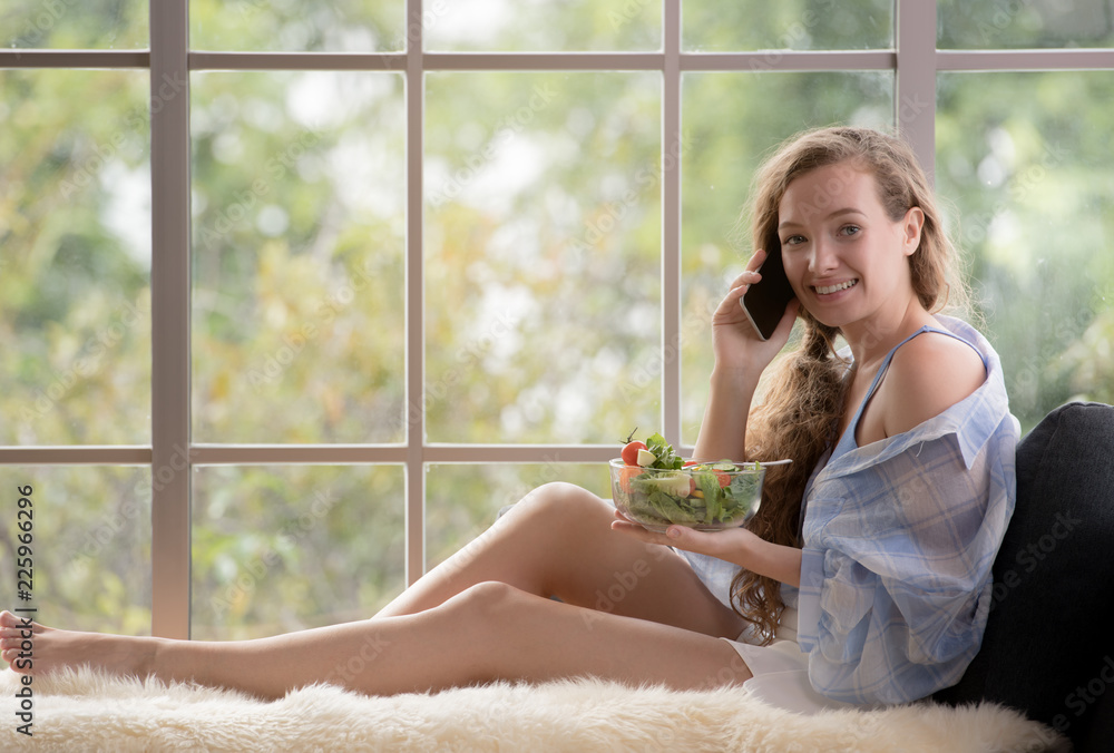 Healthy young woman lying on a couch using smartphone and holding a salad bowl looking relaxed and c