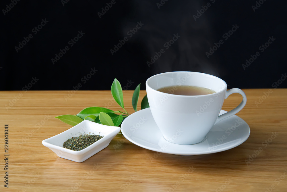 装在白色杯子里的绿茶放在一个白色茶托上，杯子上有热蒸汽。放在木桌上。