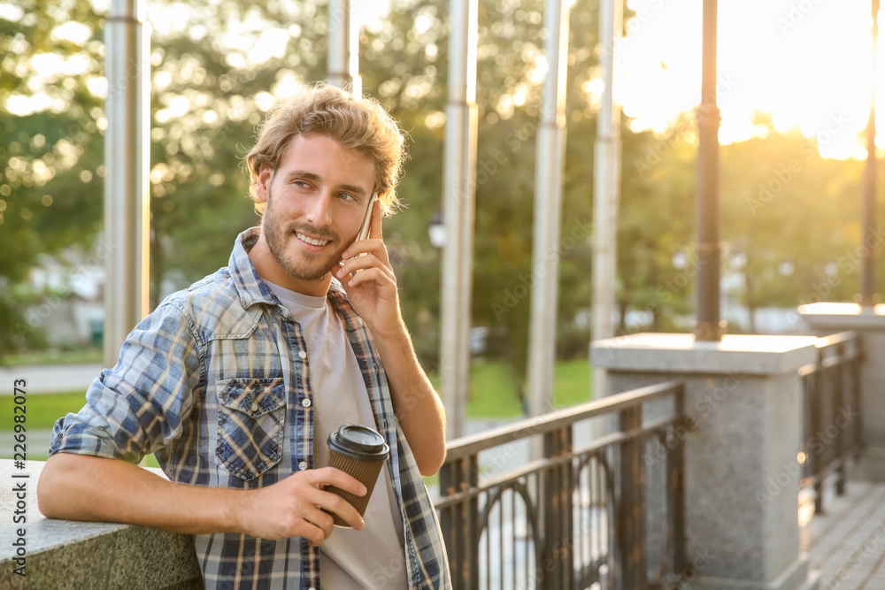 Handsome young man with coffee talking on mobile phone in park