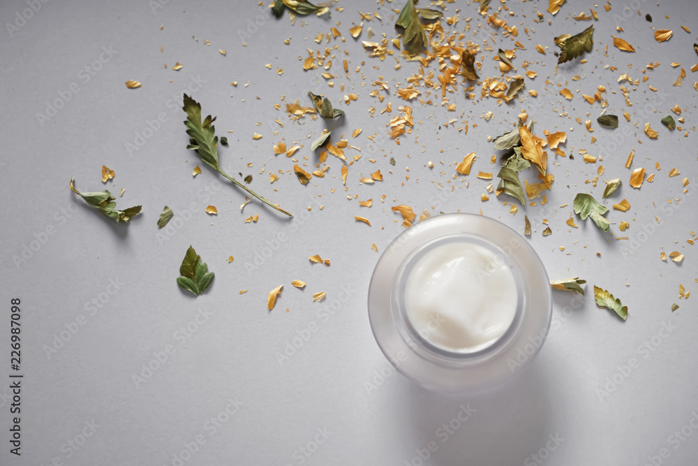 Jar with body cream and crumbled leaves on grey background