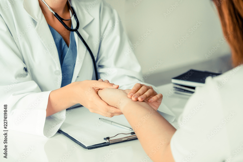 Woman doctor talks to female patient in hospital office while examining the patients pulse by hands.