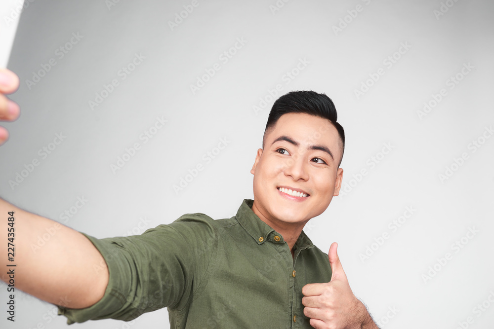 Handsome guy in white shirt is doing selfie using a smart phone, showing Ok sign and smiling, on gra