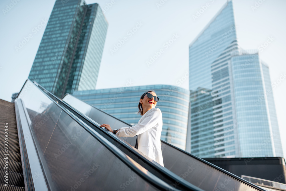 Stylish businesswoman in white suit going up on the escalator at the business centre outdoors with s