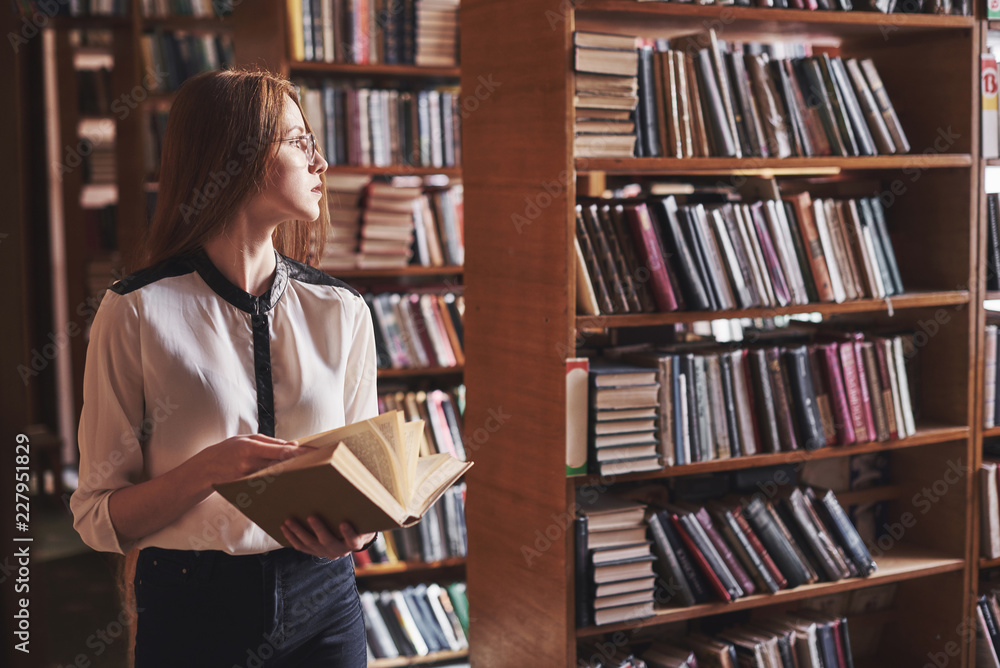 Young attractive student librarian reading a book between library bookshelves