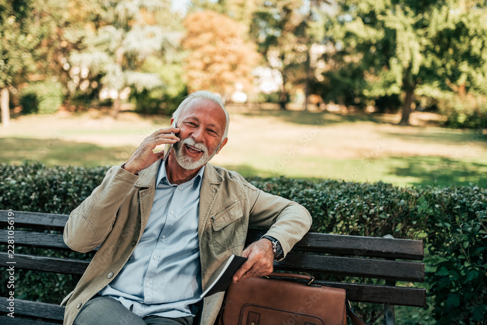 Older man talking on the phone with sitting on the bench in the park.