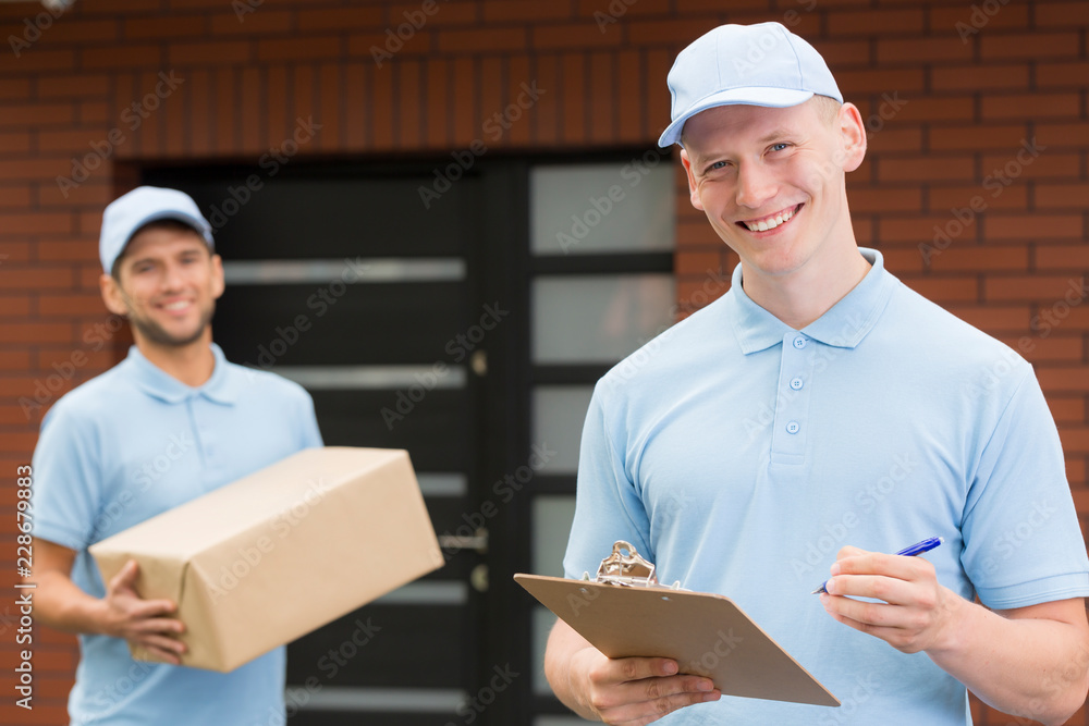 Two handsome couriers in blue uniforms standing in front of a house and waiting with delivery