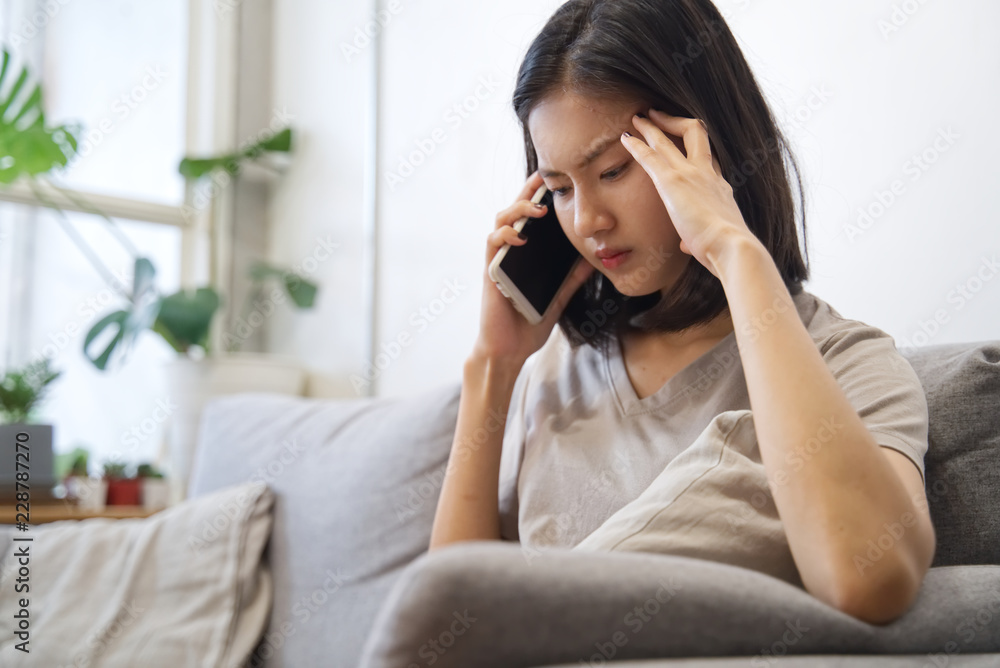 Upset/unhappy Asian girl talking on telephone is sitting on sofa at home. She feels uncomfortable an