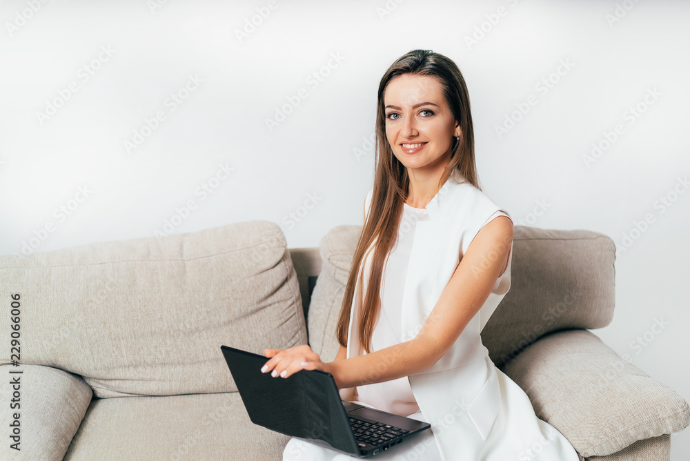 beautiful business woman in white clothes sits on a sofa and holds a laptop on her lap on a white ba