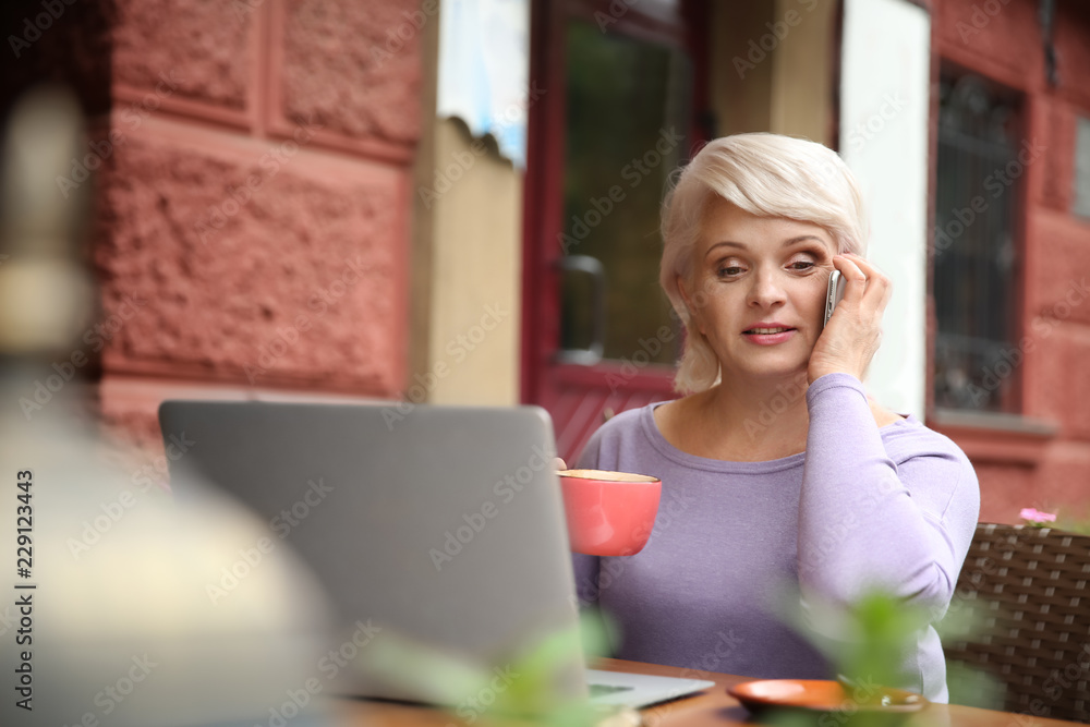 Beautiful mature woman with laptop drinking coffee and talking by phone in cafe