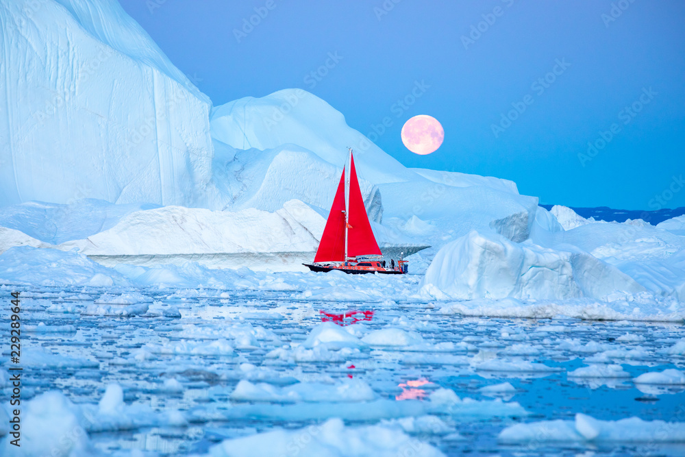 Little red sailboat cruising among floating icebergs in Disko Bay glacier during midnight sun season