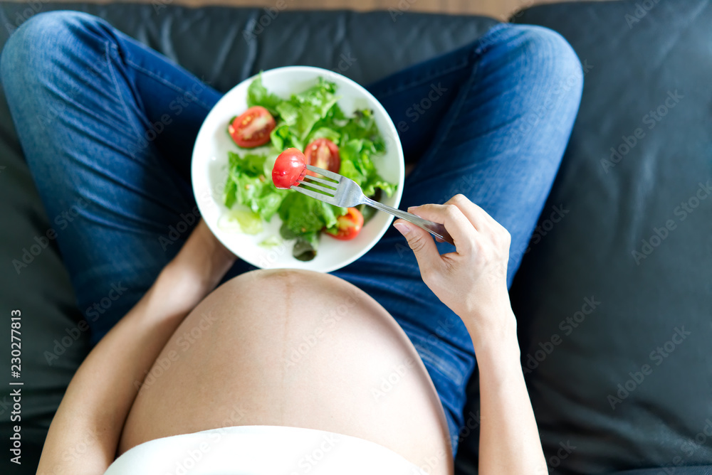 Pregnant women sitting on sofa is holding salad bowl in her left hand. She is holding fork with red 