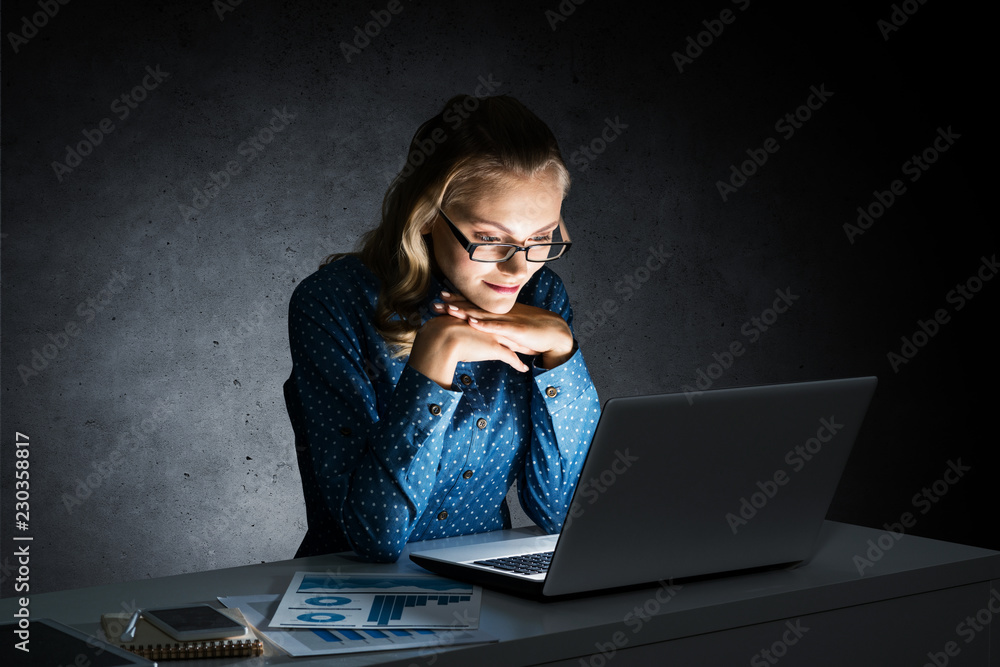 Attractive blonde working on laptop in dark office. Mixed media