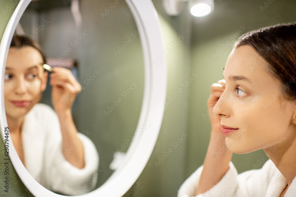 穿着浴袍的女人在浴室绿色墙上的圆镜附近画眉毛