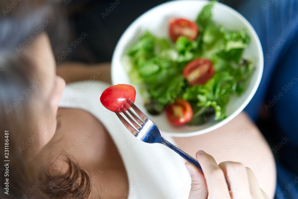 Pregnant women sitting on sofa is holding salad bowl in her left hand. She is holding fork with red 