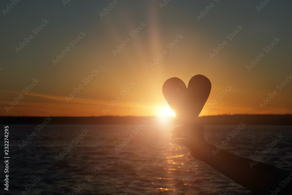 Young woman holding decorative heart on river bank at sunset