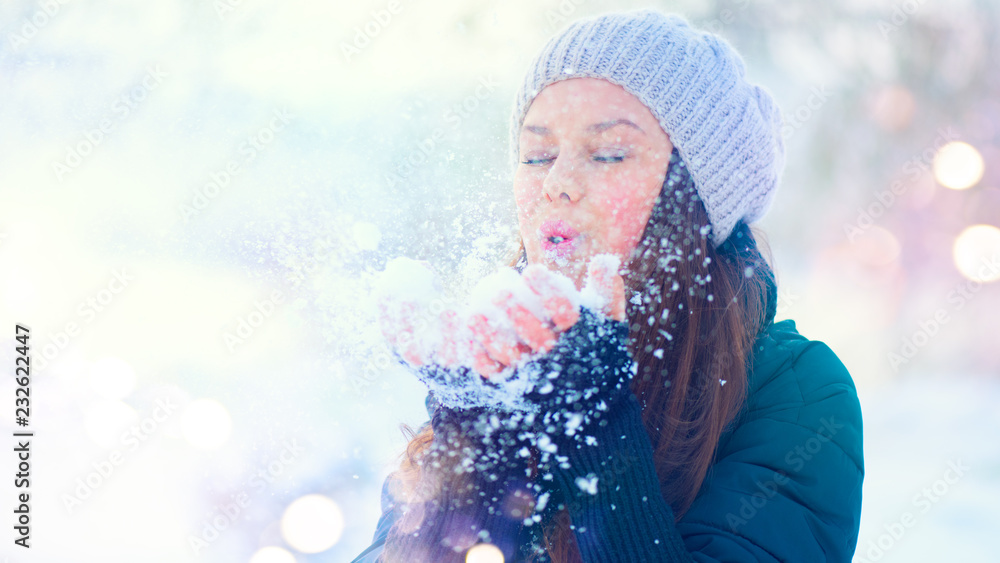 Winter girl portrait. Beauty joyful model girl blowing snow, having fun in winter park. Beautiful yo