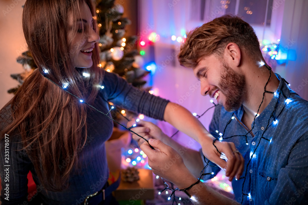 Happy couple having fun with Christmas lights next to Xmas tree at holiday evening