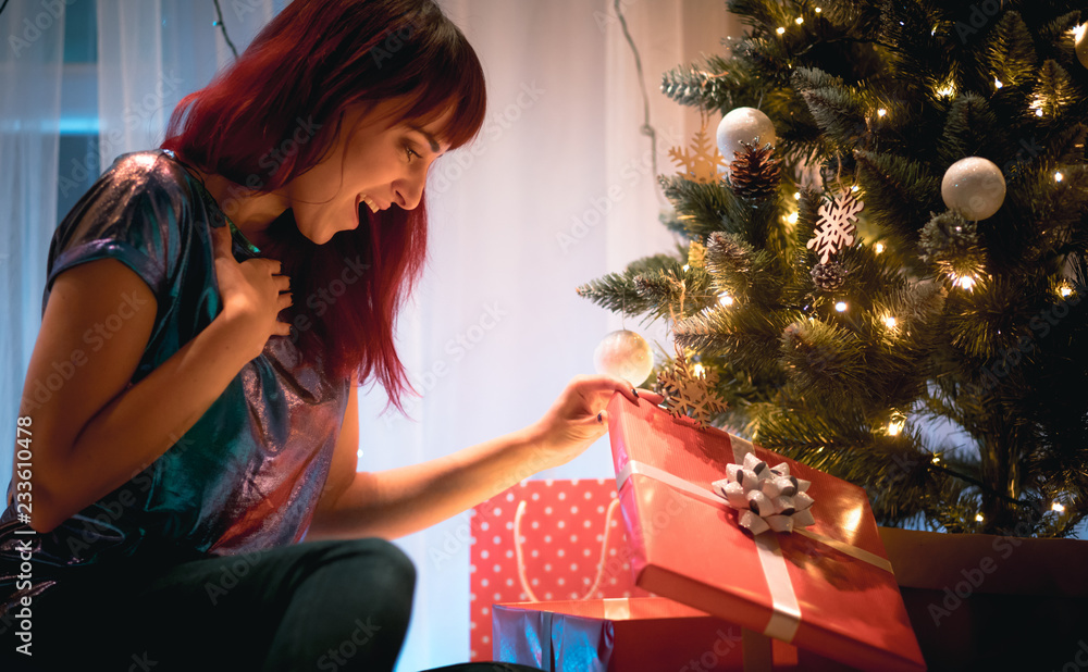 Delighted smiling woman opening Christmas gift box under tree at home