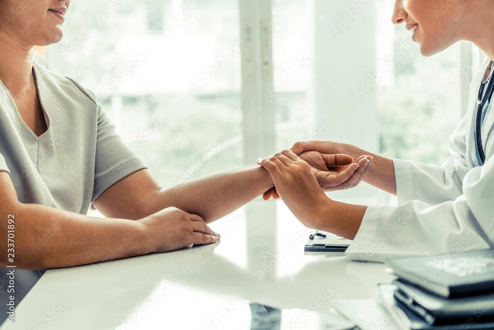 Woman doctor talks to female patient in hospital office while examining the patients pulse by hands.