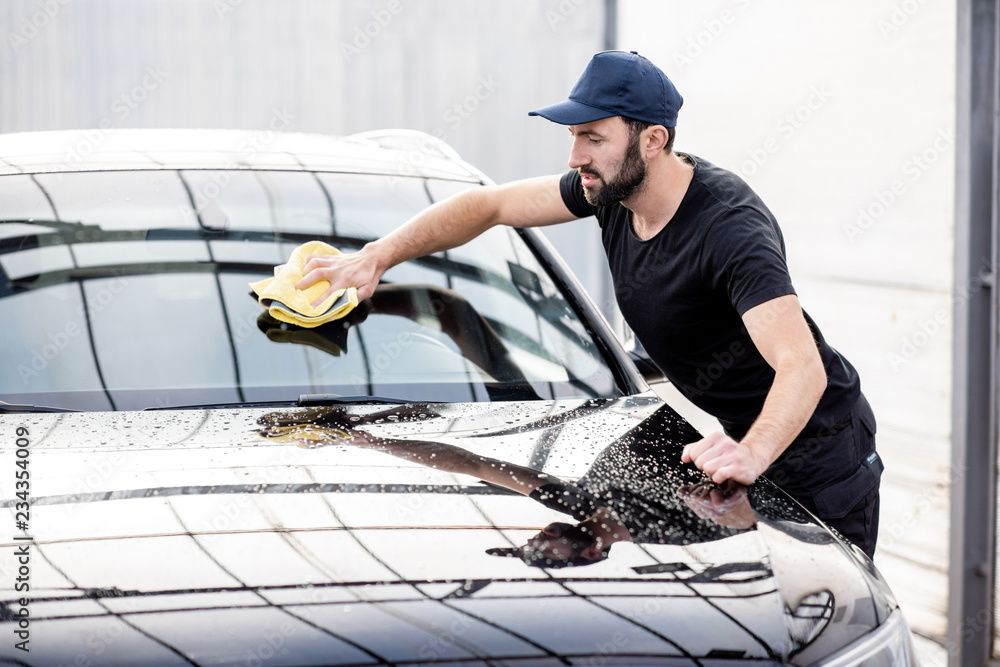 Professional washer in t-shirt and cap wiping windshield with yellow microfiber at the open air car 