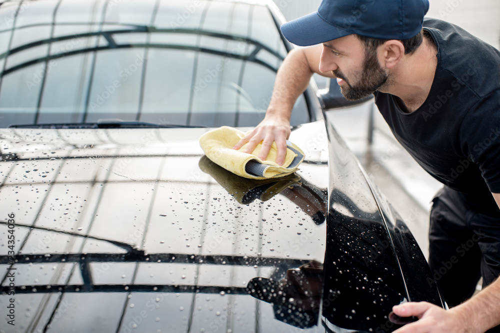 Handsome washer in t-shirt and cap wiping windshield with yellow microfiber at the open air car wash