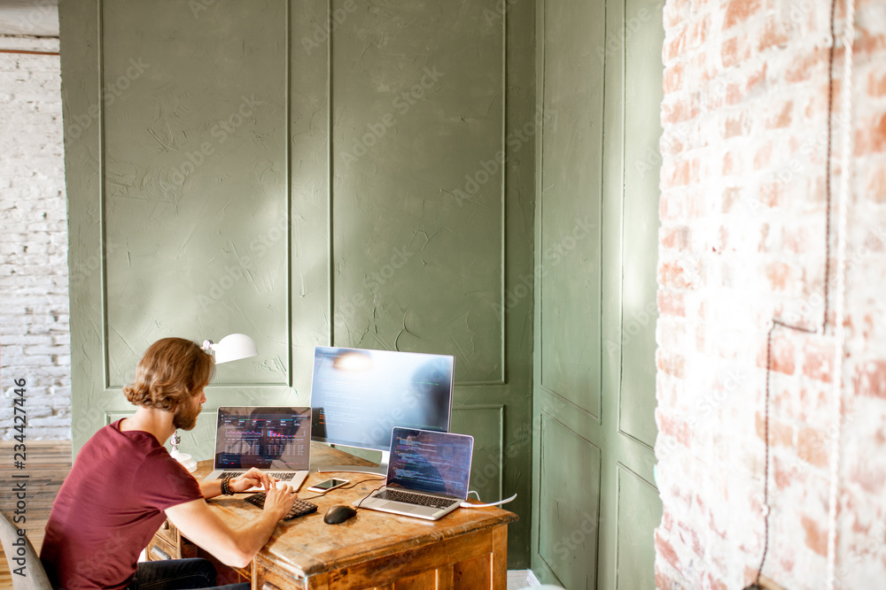 Young programmer writing a program code sitting at the workplace with three monitors in the office o