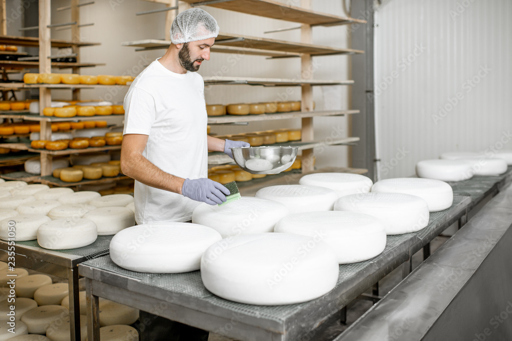 Man rubing cheese wheels with wax at the cheese manufacturing with shelves full of cheese on the bac