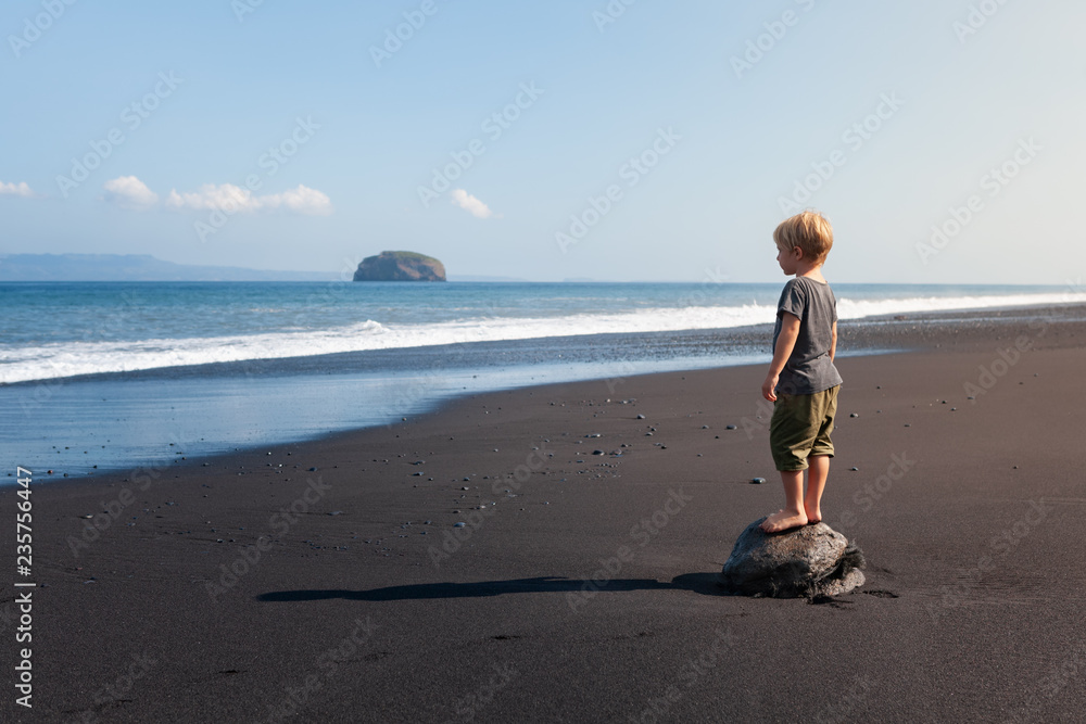 小孩站在黑沙滩上的大石头上。做梦的孩子看着海浪。孤独