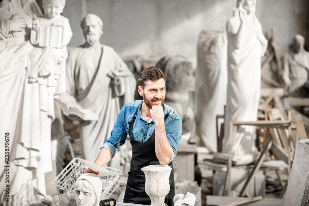 Portrait of a handsome thoughtful sculptor in blue t-shirt and apron working with stone sculptures o