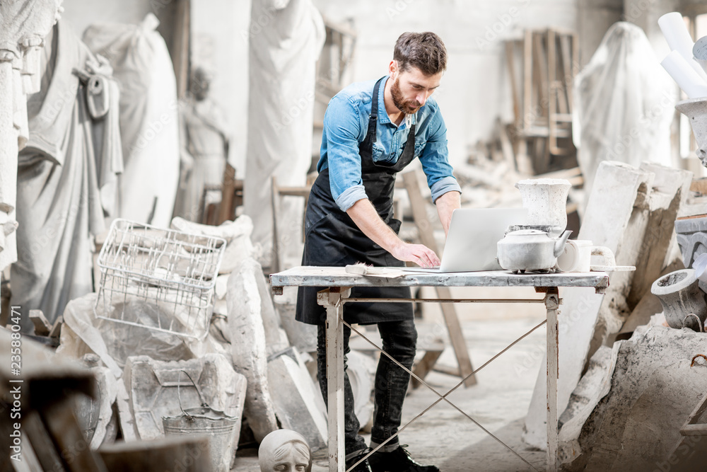 Handsome sculptor working with laptop at the working place in the old studio with sculptures on the 